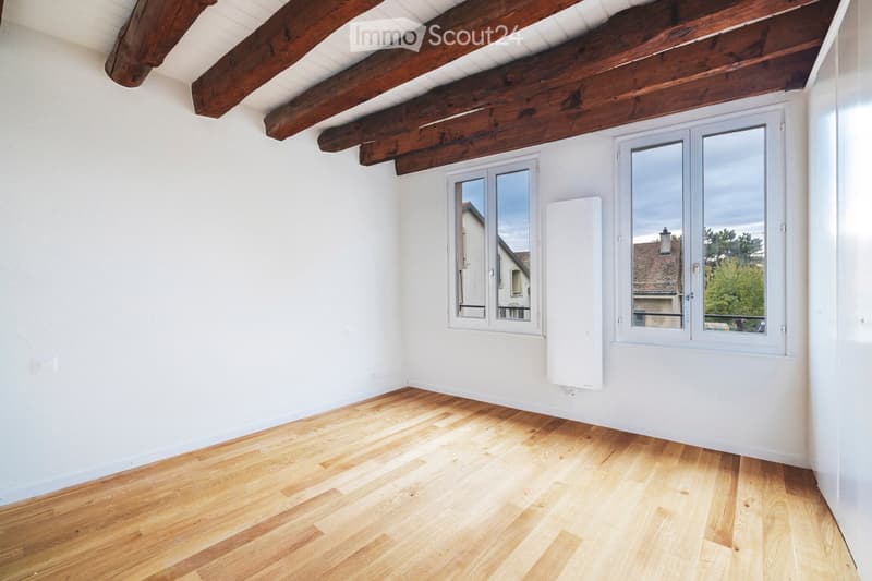 Empty room with wooden flooring, white walls, and exposed wooden beams on the ceiling. Two large windows with white frames offer natural light and views of the outside.