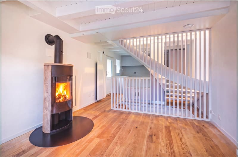 Modern hallway with wooden flooring, white walls, and a contemporary fireplace with a visible flame. A white staircase with a sleek railing is visible in the background.