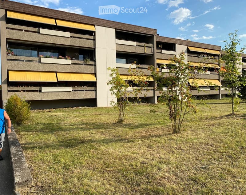 Multi-story residential building with balconies, yellow awnings, and surrounding greenery.