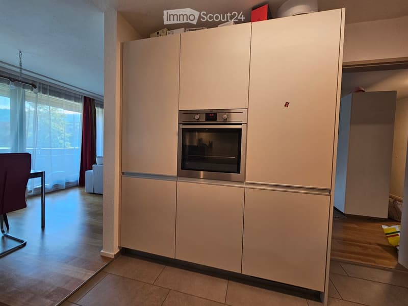 Modern kitchen with white cabinets, stainless steel oven, refrigerator, and a small seating area with a chair and table near a large window with curtains.