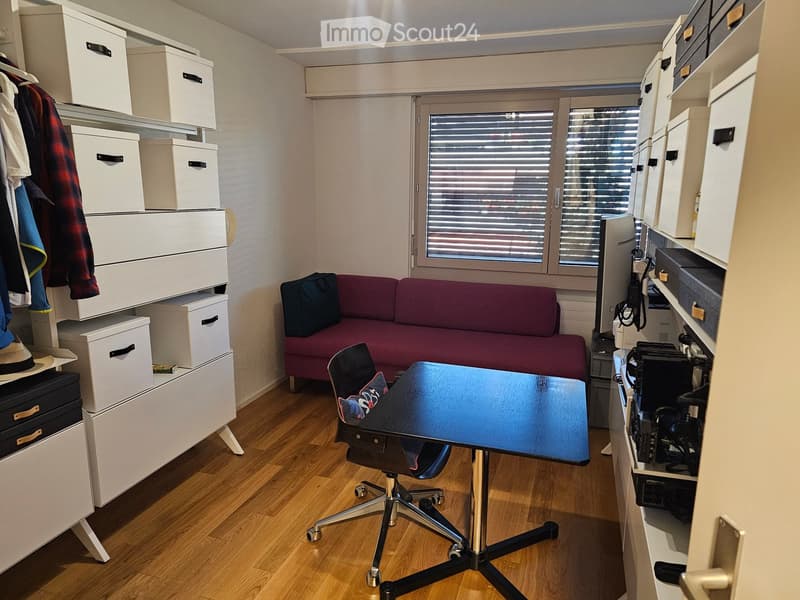 A compact, furnished room with a red sofa, a blue table, a black office chair, and multiple storage units with boxes and a hat on top.
