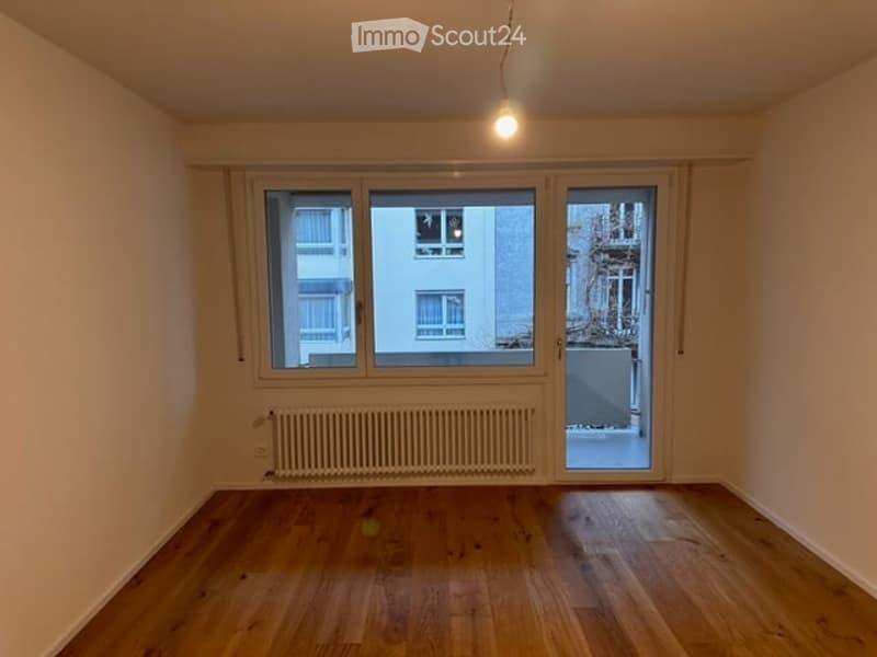 Empty room with wooden flooring, large windows, and a radiator. No furniture or equipment present.