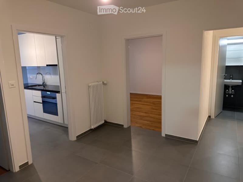 Empty room with grey tiled floor, white walls, radiator, and open doorways to a kitchen and bathroom.