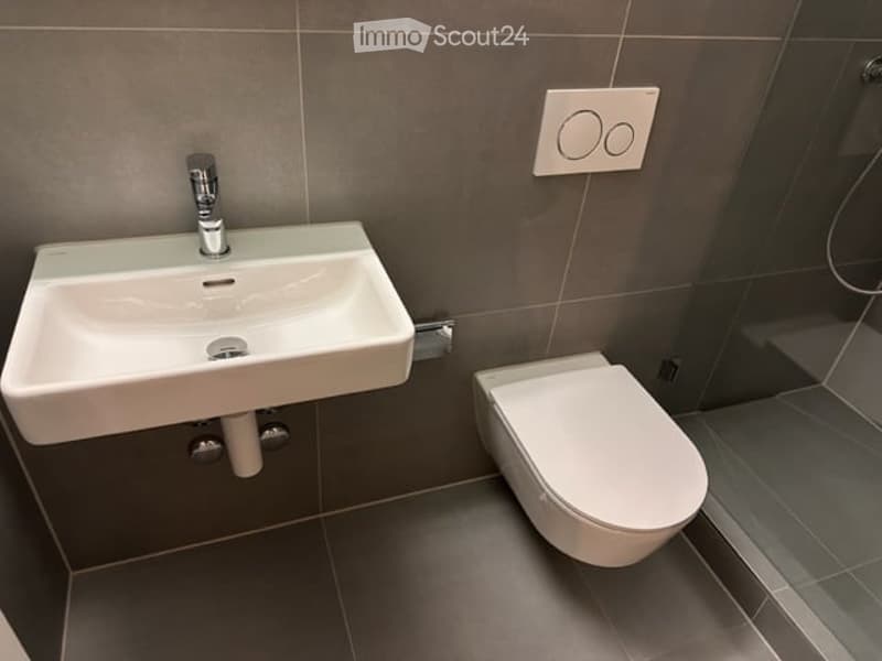 Modern bathroom with a white sink, chrome faucet, and white toilet. Grey tiled walls and floor.