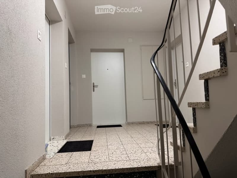 A clean, modern hallway with tiled flooring, a closed door at the end, and a staircase with a metal railing on the right side.