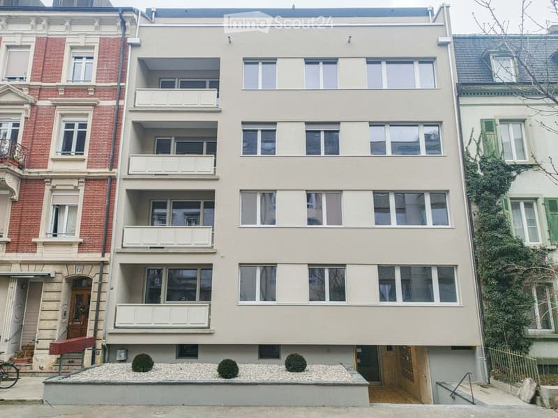 Four-story residential building with beige facade, multiple windows, and white balconies. Adjacent buildings include red brick and green-shuttered structures. Small landscaped area with gravel and shrubs in front.