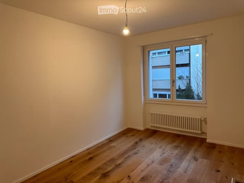 Empty room with wooden flooring, white walls, a single window with a view of neighboring buildings, and a radiator below the window.