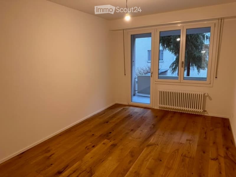 Empty room with wooden flooring, large window with sliding glass door leading to a balcony, radiator, and a single hanging light fixture.