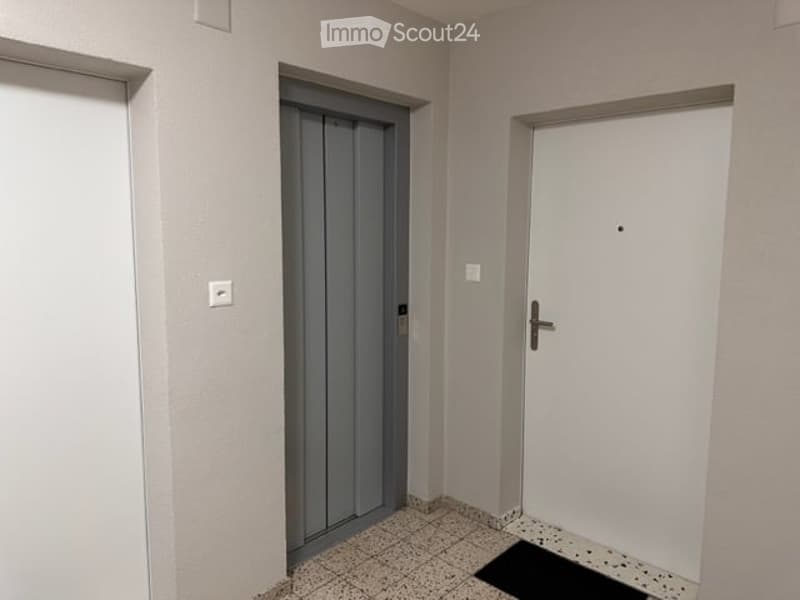 Empty hallway with tiled floor, white walls, and two doors: one grey elevator door and one white door.