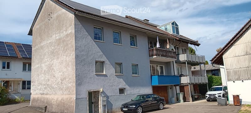 Multi-story residential building with balconies, solar panels on the roof, and a parking area with several cars.