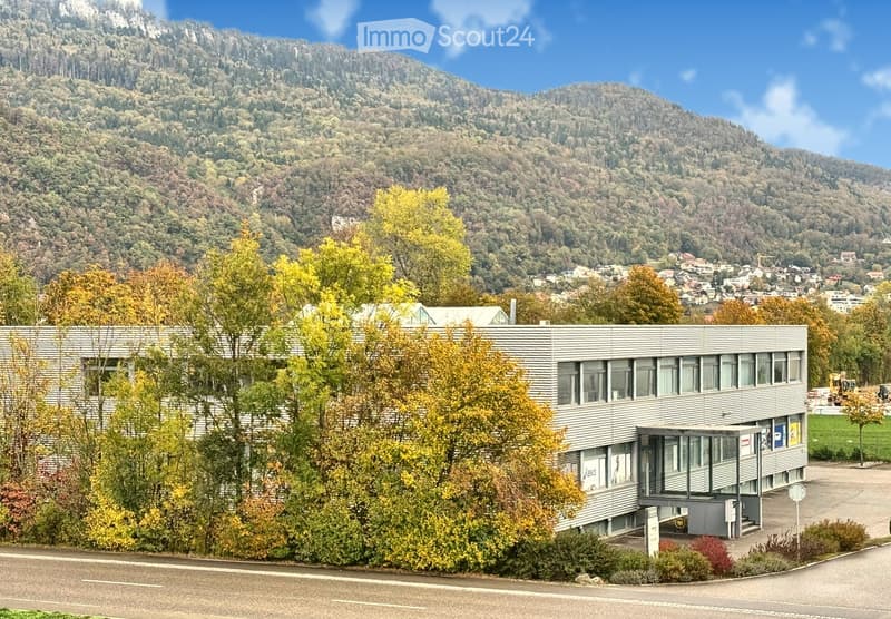 Modern, two-story building with a grey metal facade and large windows, surrounded by autumn trees and situated in a scenic valley with mountains in the background.