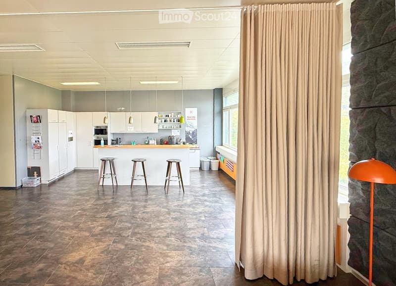 Modern kitchen area with white cabinets, a central island, three stools, and a refrigerator. The space features a tiled floor and large windows with curtains.