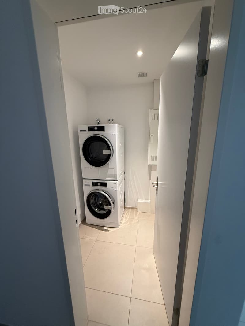 A small, well-lit room with white tiled flooring and walls, containing a stacked washing machine and dryer unit.