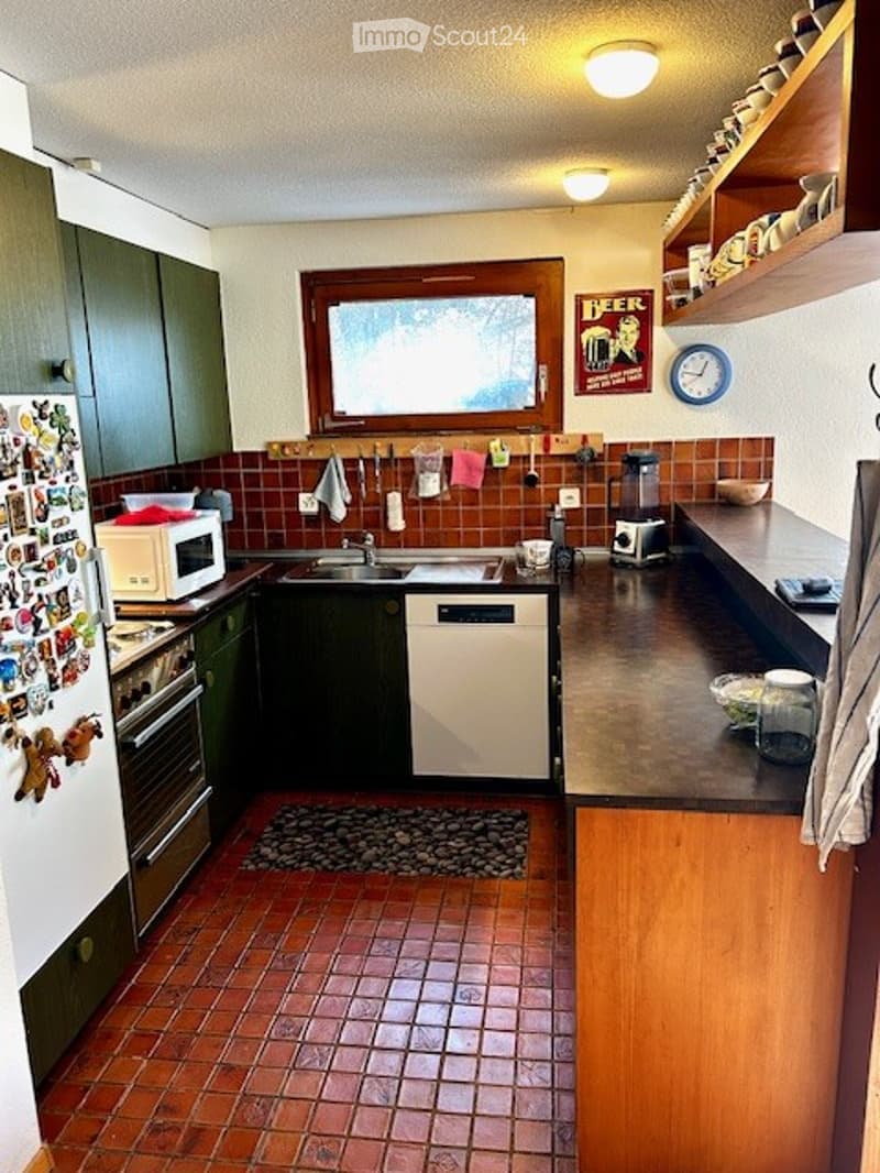 A compact kitchen with dark green cabinets, a red brick backsplash, and a tiled floor. Features include a refrigerator, microwave, oven, dishwasher, sink, blender, and a clock on the wall.