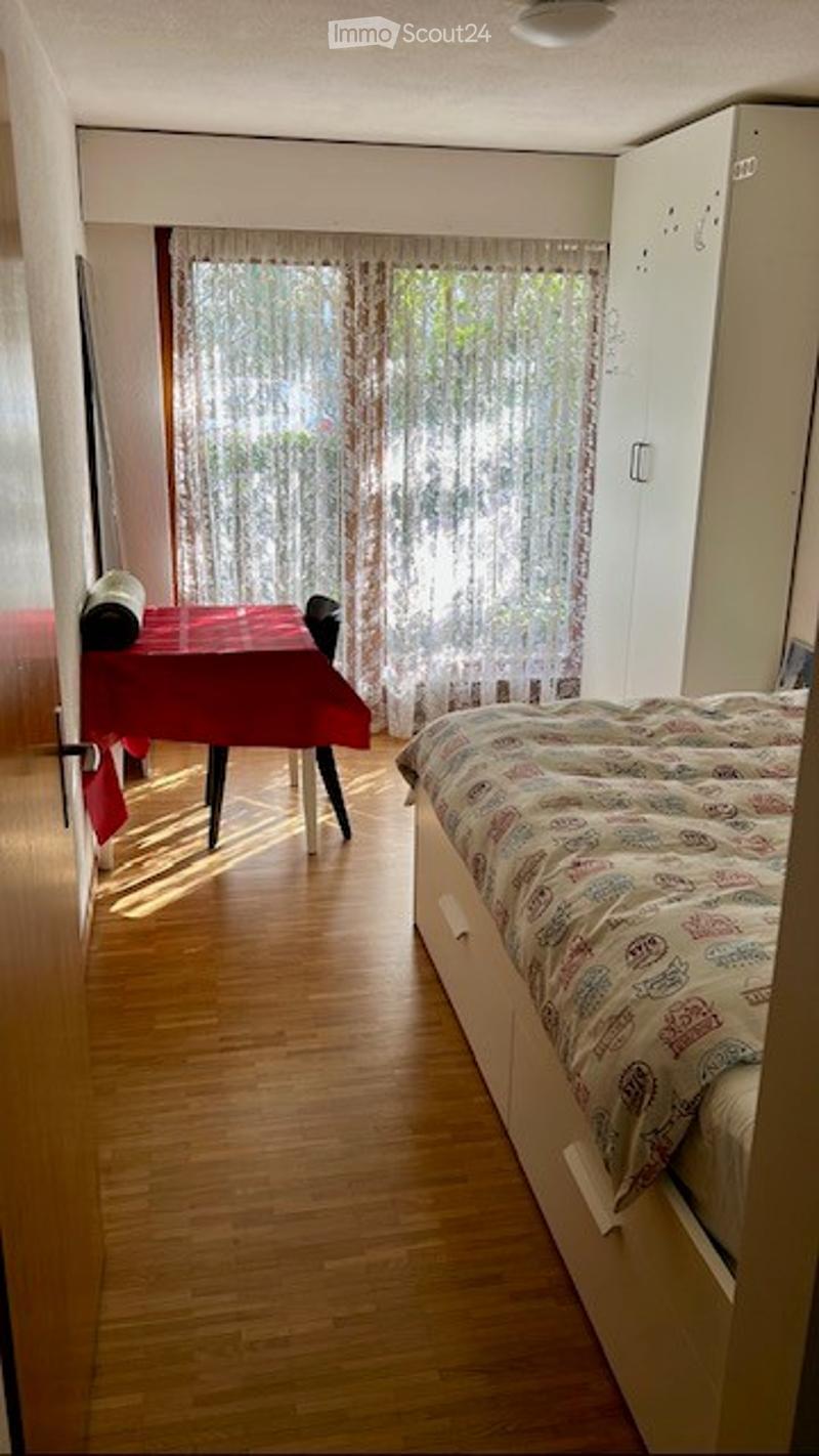 Bright bedroom with wooden flooring, a large bed with patterned bedding, a white wardrobe, and a small dining area with a red tablecloth and chairs.