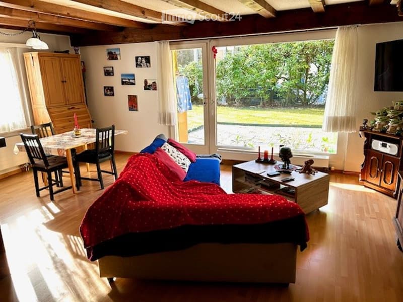Bright living room with wooden flooring, large windows offering a view of the garden, a red blanket-covered sofa, a coffee table, a dining area with a table and chairs, and a wooden cabinet.