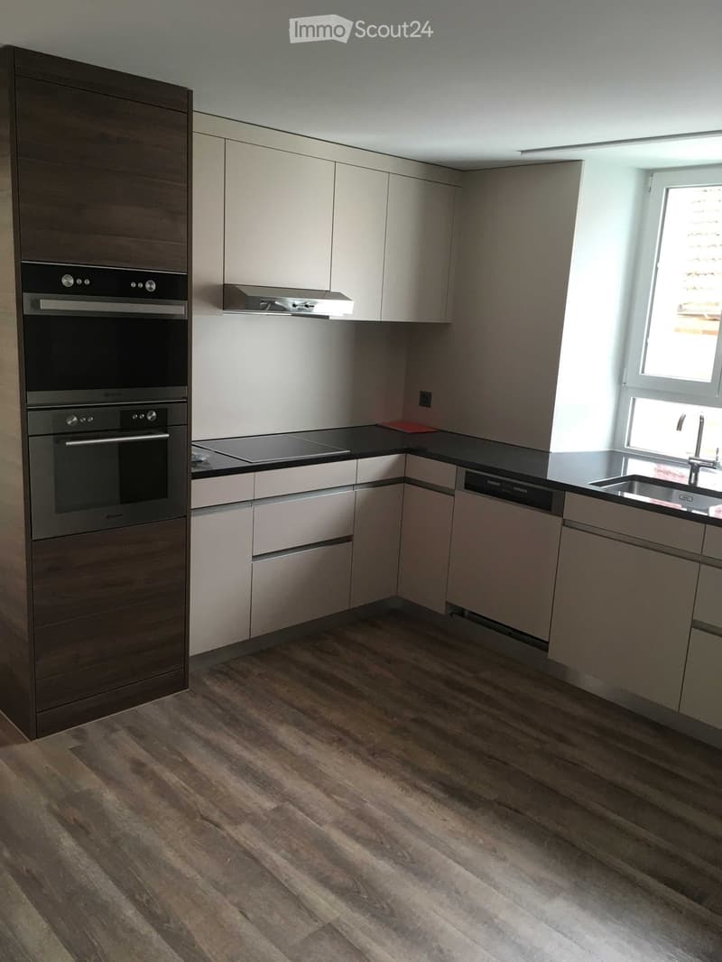 Modern kitchen with light-colored cabinets, dark countertops, and wooden flooring. Features an oven, dishwasher, and sink with a faucet.