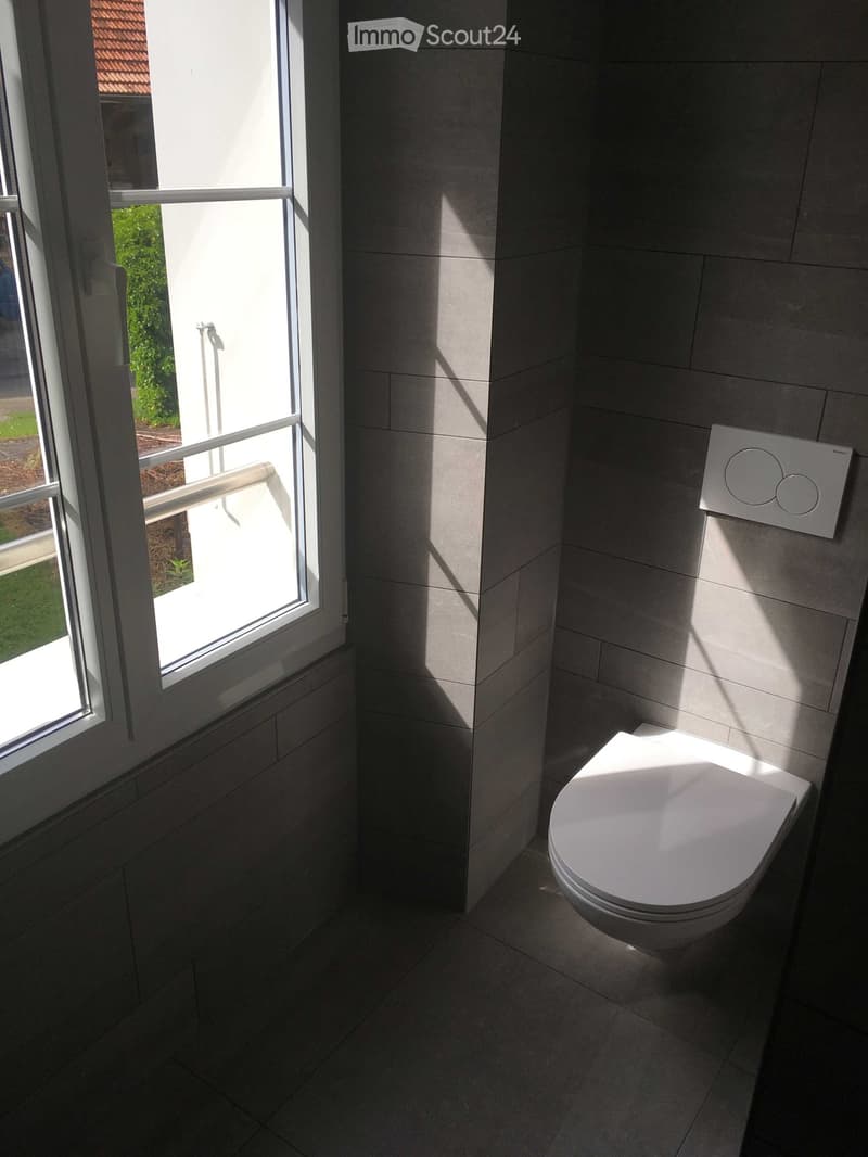 Modern bathroom with a toilet, control panel, and large window with a view of greenery outside.