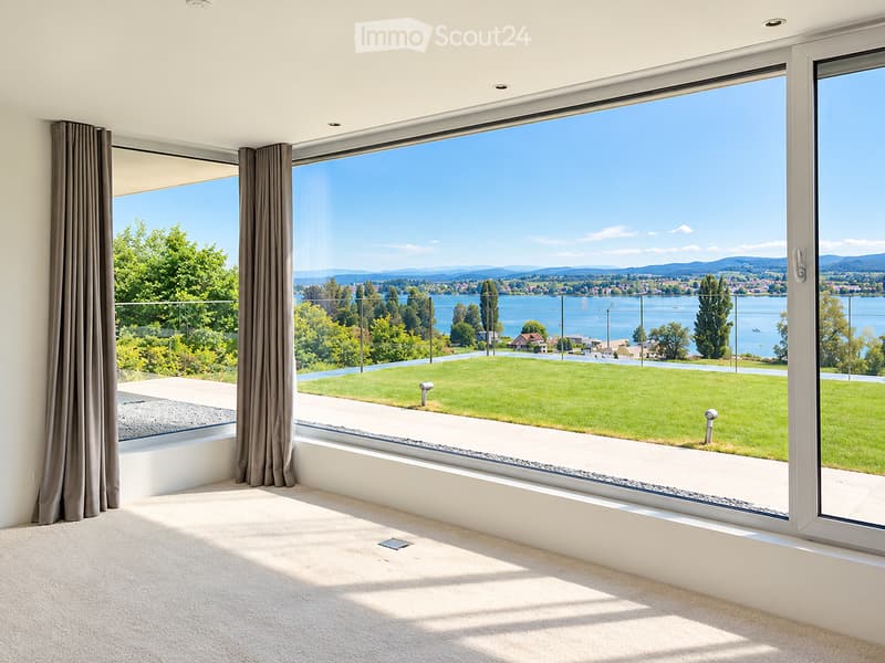 Bright, unfurnished room with large windows offering a scenic view of a lake and surrounding landscape. The room features carpeted flooring and modern window frames.