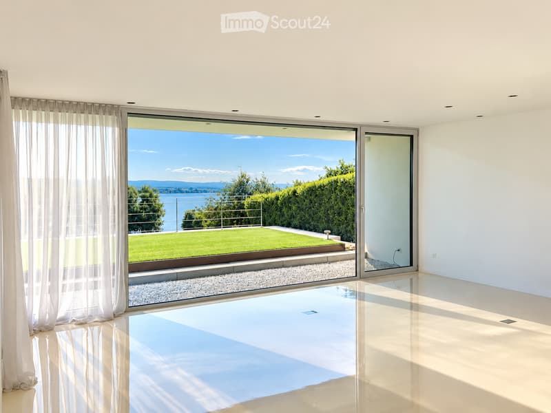 Bright, unfurnished room with large glass doors leading to a view of a green lawn and water. The floor is polished concrete, and the walls are white.