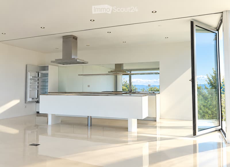 Modern, minimalist kitchen with white cabinetry, a central island, and a large range hood. The space features large windows offering a scenic view of the outdoors.