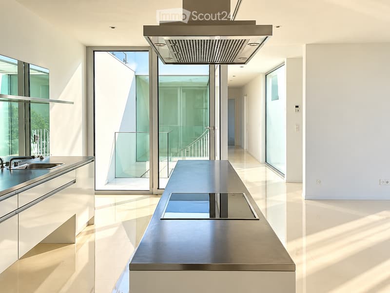 Modern kitchen with sleek white cabinets, a central island, and a built-in range hood. Large glass doors open to a bright outdoor area.