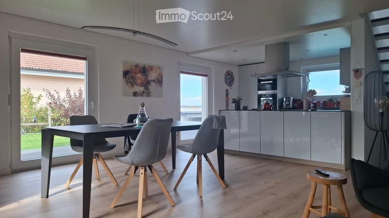 Open-plan dining and kitchen area with modern furniture, including a dining table with chairs, a small stool, and a fitted kitchen with cabinets and appliances.