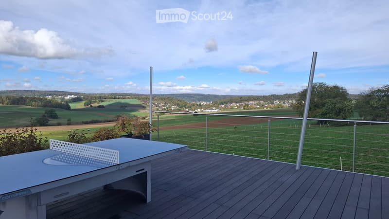 A modern balcony with a ping pong table, wooden decking, and a metal railing overlooking a scenic countryside view.