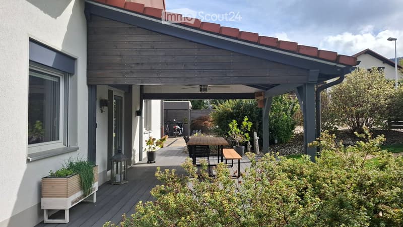 A covered patio area with wooden decking, a table and chairs, a bench, lantern, and potted plants.