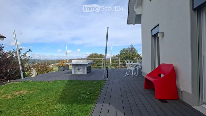 Balcony with wooden decking, modern furniture including a red chair, white table, and white chairs, overlooking a scenic landscape.