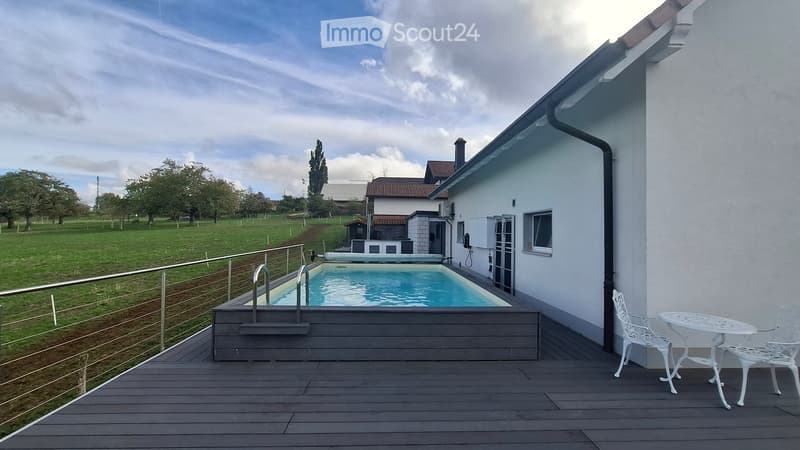 Outdoor patio with a swimming pool, wooden decking, white metal table and chairs, and a view of a grassy field and trees.