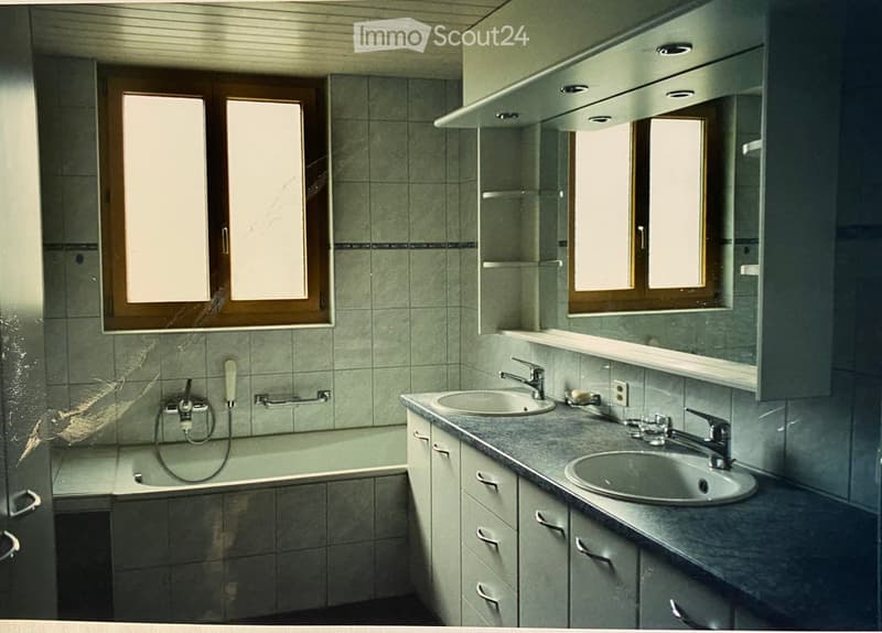 Bathroom with a bathtub, two sinks, cabinets, and windows. The walls are tiled, and the countertop is made of dark stone.