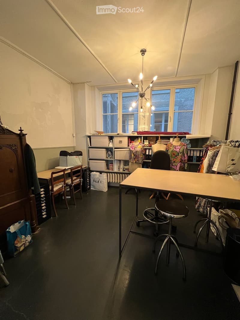 A well-lit room with large windows, a wooden table, a chair, a stool, a wardrobe, shelves with storage boxes, mannequins, and a radiator.