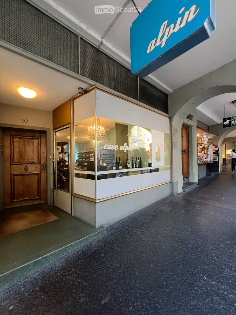 A well-lit hallway with a shop entrance labeled "cave alpine" featuring glass display cases with bottles, a chandelier inside, and a wooden door to the left.