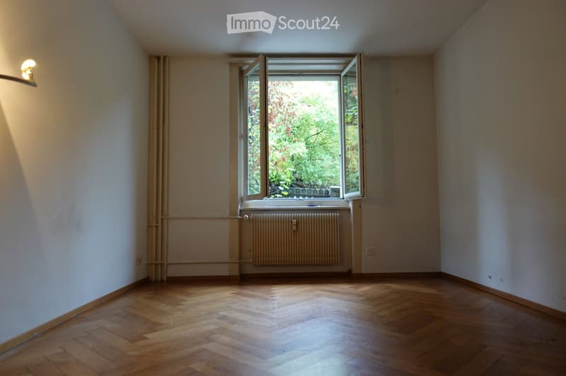 Empty room with wooden flooring, white walls, large window with open shutters, radiator, and a wall-mounted light fixture.