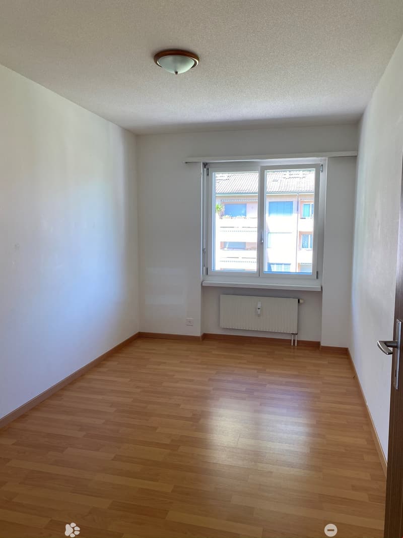 Empty room with wooden flooring, white walls, a large window with blinds, a radiator, and a ceiling light fixture.