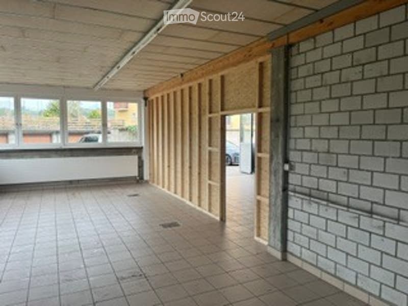 Empty room with tiled floor, exposed wooden beams, and large windows, showing unfinished walls and a doorway leading outside.