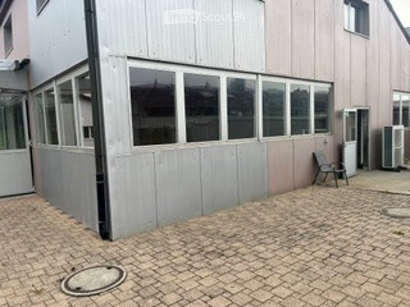 Brick-paved outdoor area adjacent to a building with large windows and a grey panel wall. A single chair is placed near the entrance.