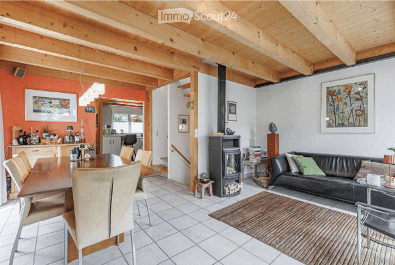 Bright, open-plan living and dining area with exposed wooden beams, a black leather sofa, a wooden dining table with chairs, a fireplace, and a small kitchen area visible in the background.