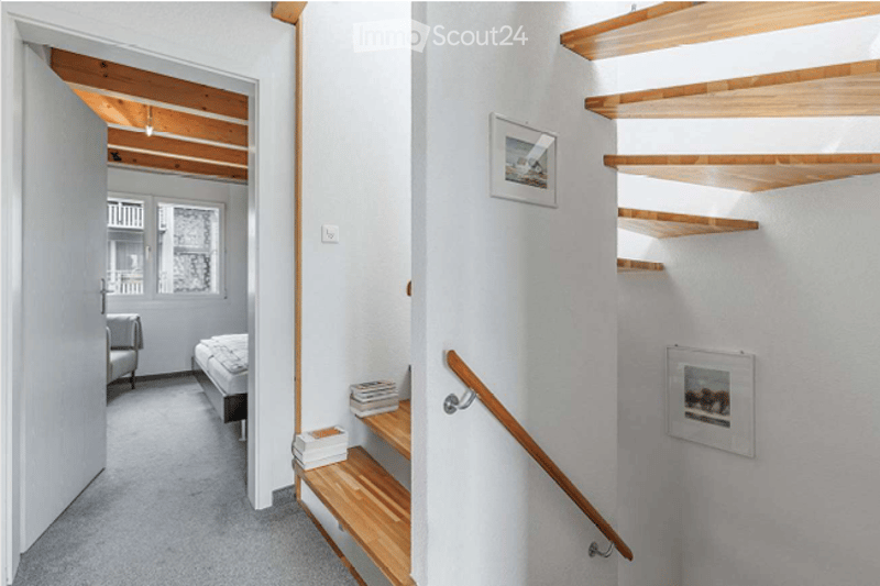 A modern hallway with a wooden staircase, white walls, and a few framed pictures. The hallway leads to a bedroom with a bed and a chair visible in the background.