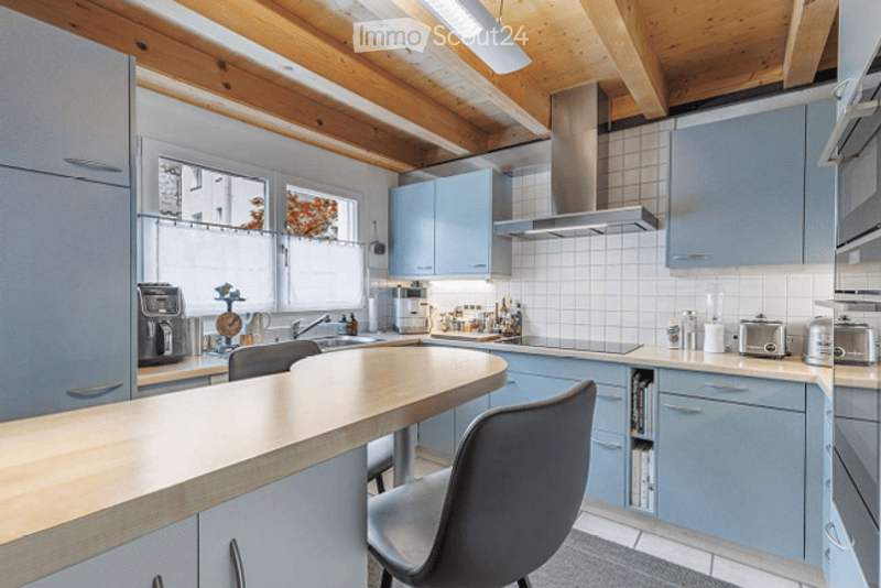 Modern kitchen with light blue cabinets, stainless steel appliances, wooden ceiling beams, and a central island with seating.