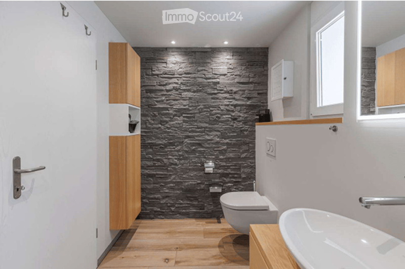 Modern bathroom with a dark stone accent wall, light wooden flooring, and white fixtures including a toilet, sink, and cabinet.