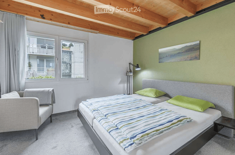 Bright bedroom with wooden ceiling beams, large windows, a bed with striped bedding, a grey armchair, and a wall-mounted lamp.