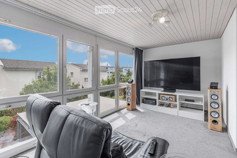 Modern living room with large windows offering a view, a grey sofa, a coffee table, a TV stand with speakers, and a remote control on the sofa.