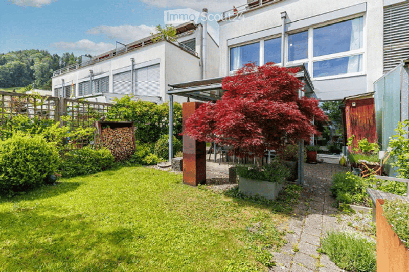 Well-maintained garden with a lush green lawn, trimmed hedges, a red maple tree, patio furniture, and a modern building in the background.