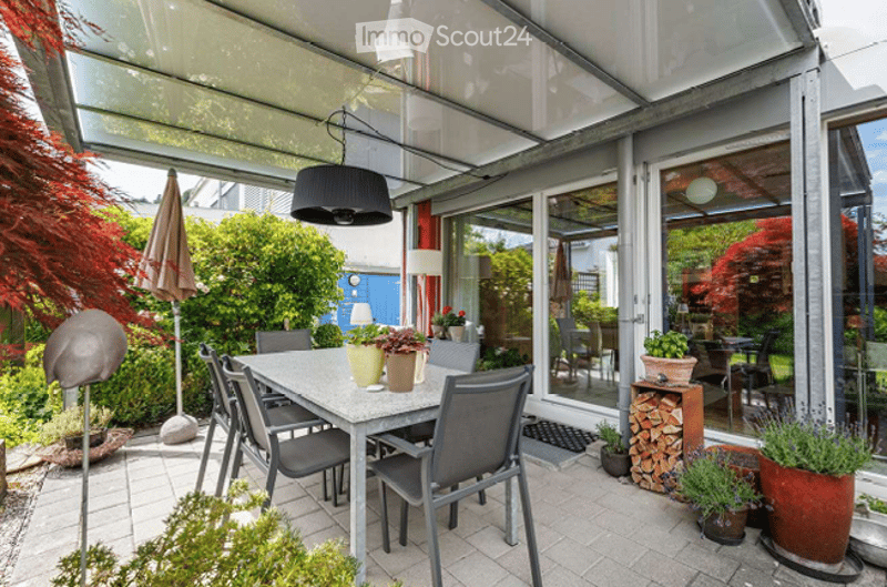 A modern, covered outdoor dining area with a glass roof, surrounded by greenery and potted plants. A dining table with chairs is set up, and there is a hanging lamp above. The space features a brick patio and is enclosed by large glass doors leading to an