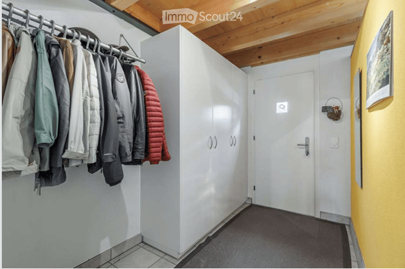 A narrow hallway with white walls, a wooden ceiling, and a coat rack holding several jackets. A closed white door is visible at the end of the hallway.