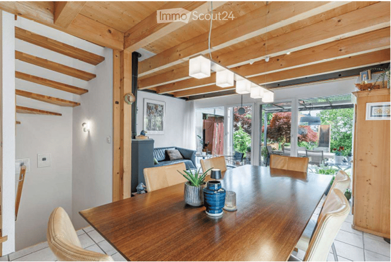 Bright, open-plan dining and living area with wooden beams, white walls, and large windows leading to a patio. Features a wooden dining table with chairs, a sofa, and decorative items on the table.