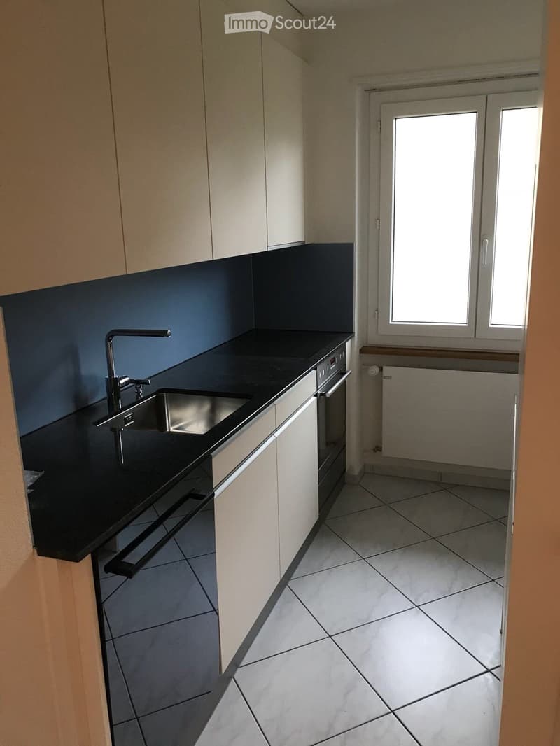Empty kitchen with white cabinets, black countertop, stainless steel sink, and tiled floor. No appliances or furniture present.