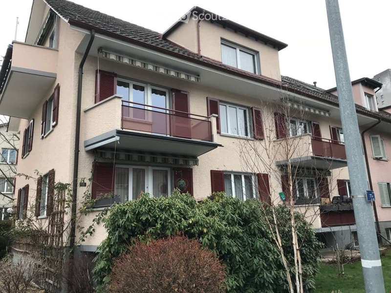 A three-story residential building with beige walls, red shutters, multiple windows, and balconies. The ground floor has a small garden area with bushes and plants.